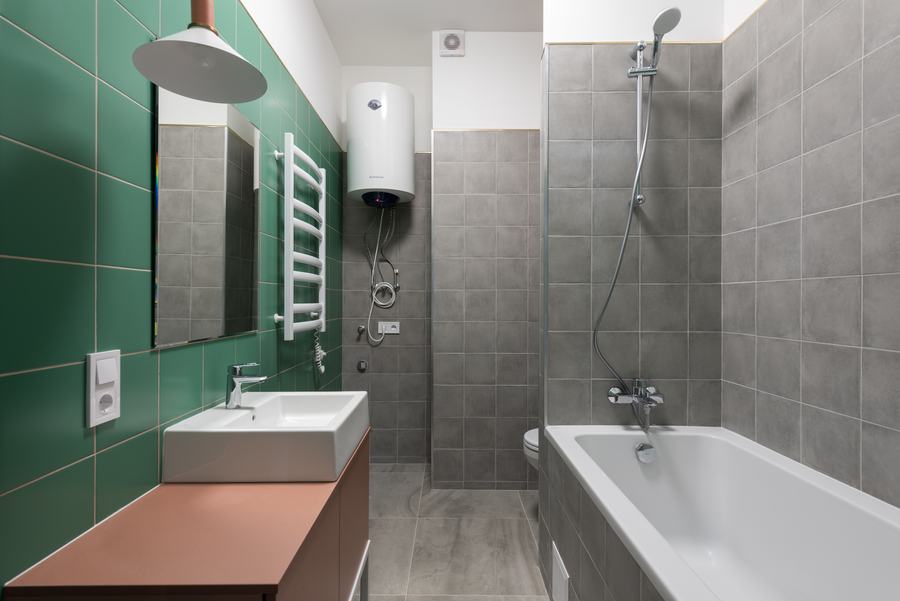 Salle de bain carrelée de vert et de gris avec un ballon d'eau chaude blanc fixé au mur au-dessus d'une baignoire